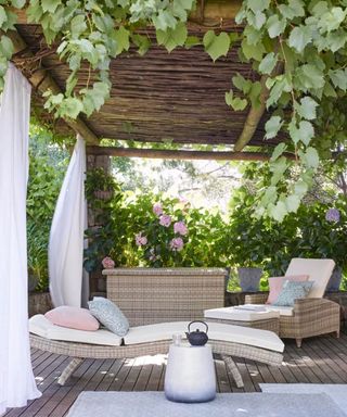 A dark brown backyard area with a light brown sunlounger and chair with blue and pink throw pillows, a rattan storage basket, a white stool with a black tea pot, and a pergola above it with green leaves