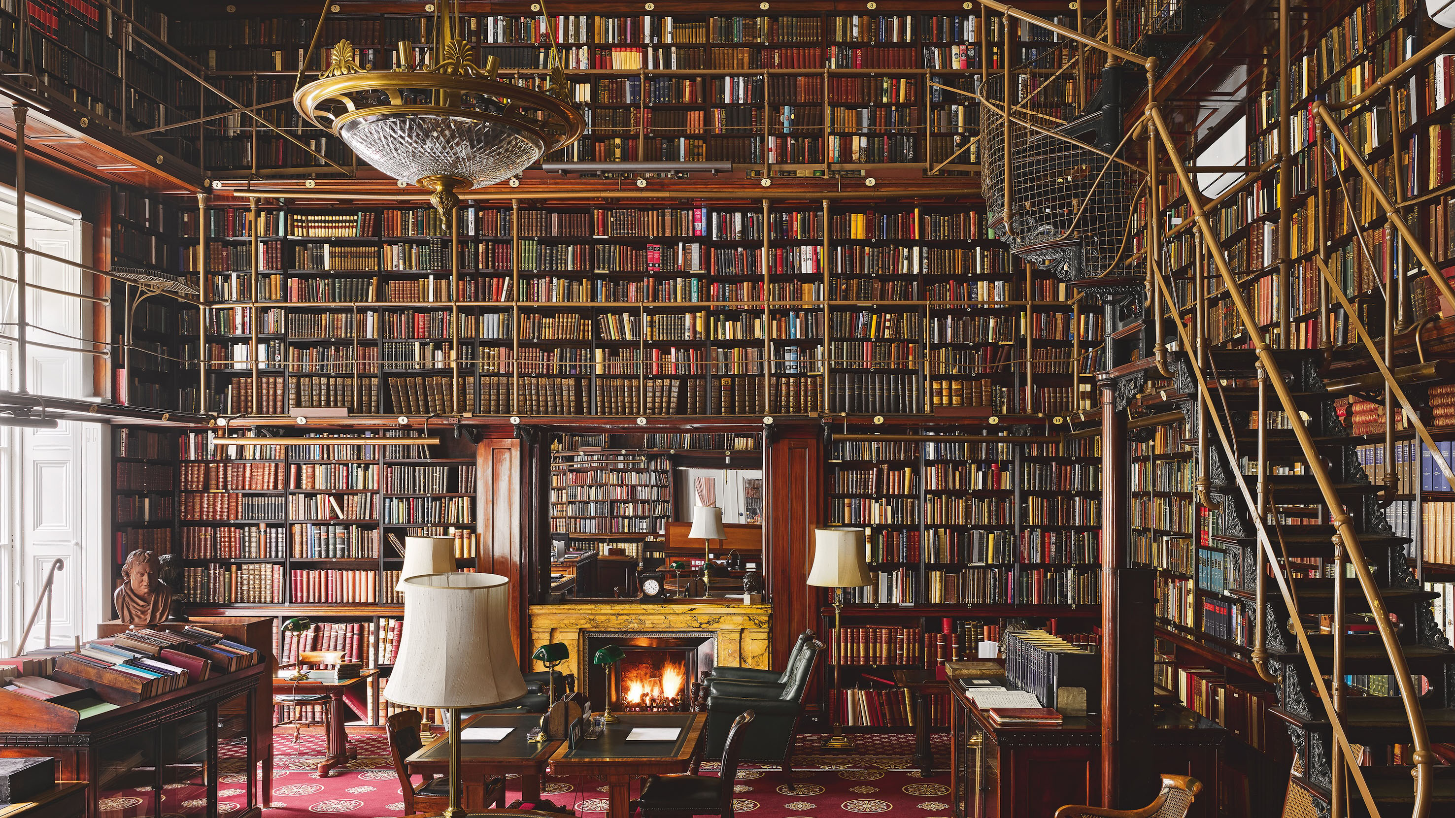 The Athenaeum library with double height shelves, a gold ladder and walkway and antique furniture on a red patterned carpet