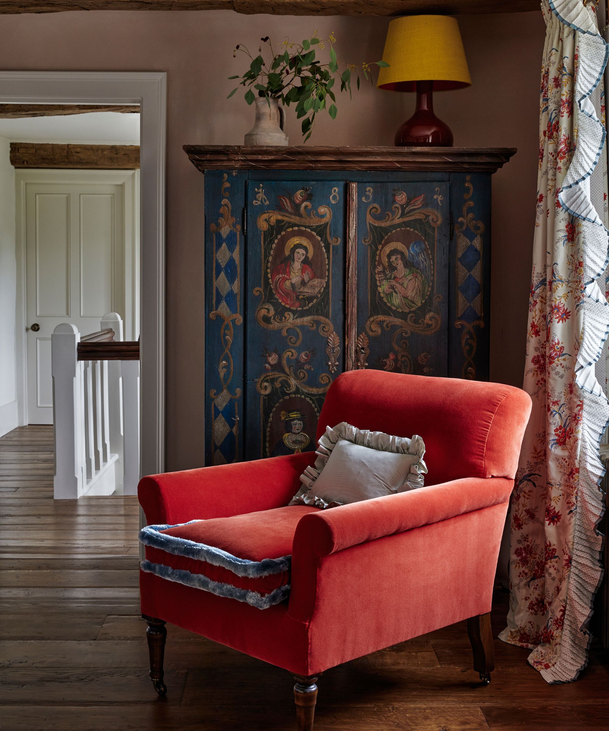 Living room with red velvet armchair, antique painted cabinet, yellow lamp and frilly curtains