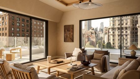 A neutral living room in a penthouse with a balcony running around it with a glass balustrade with two matching wood coffee tables in the center of mirroring armchairs and a sofa