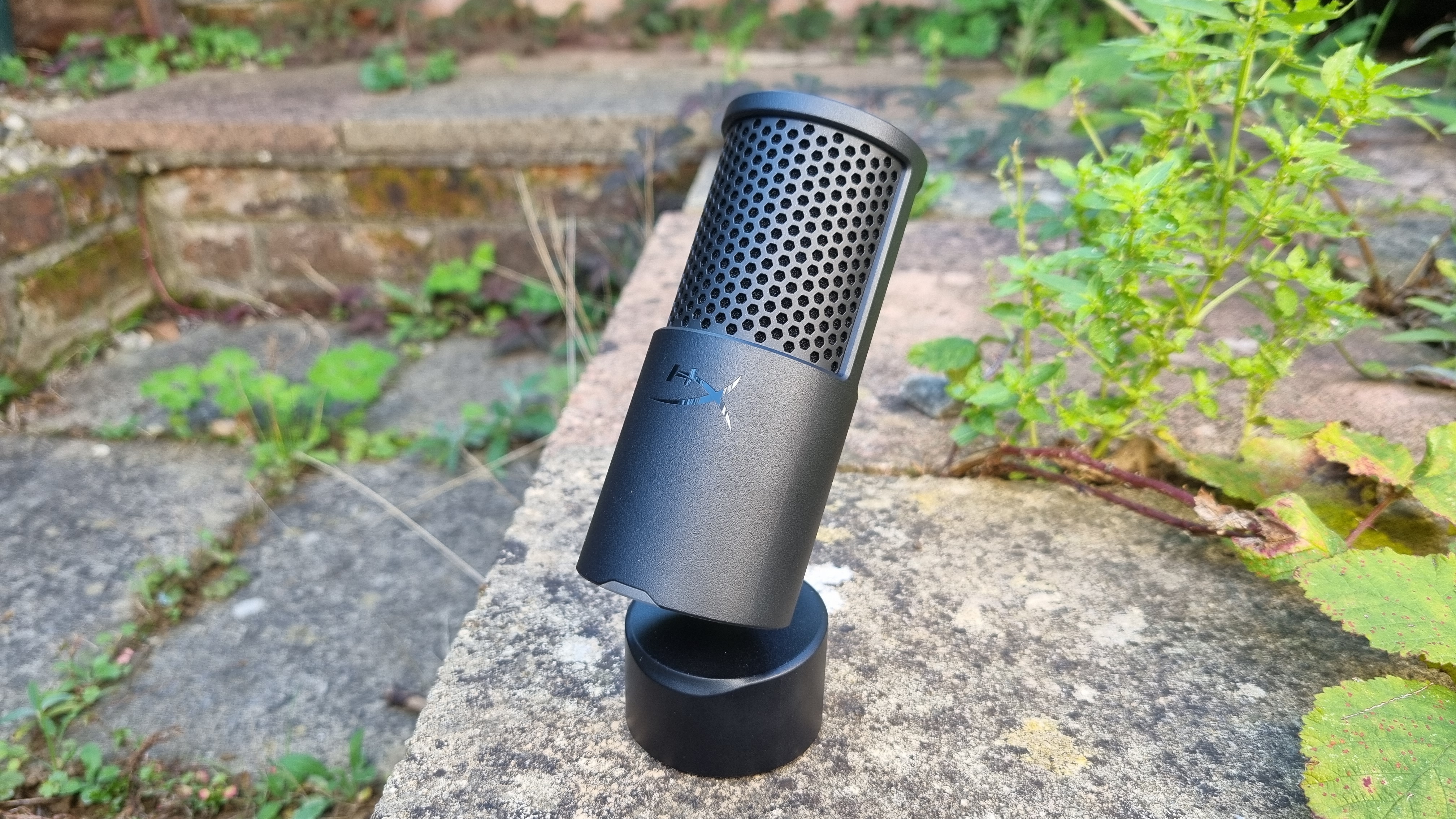 The HyperX SoloCast 2 USB microphone outside amid some foliage, with its base tilted