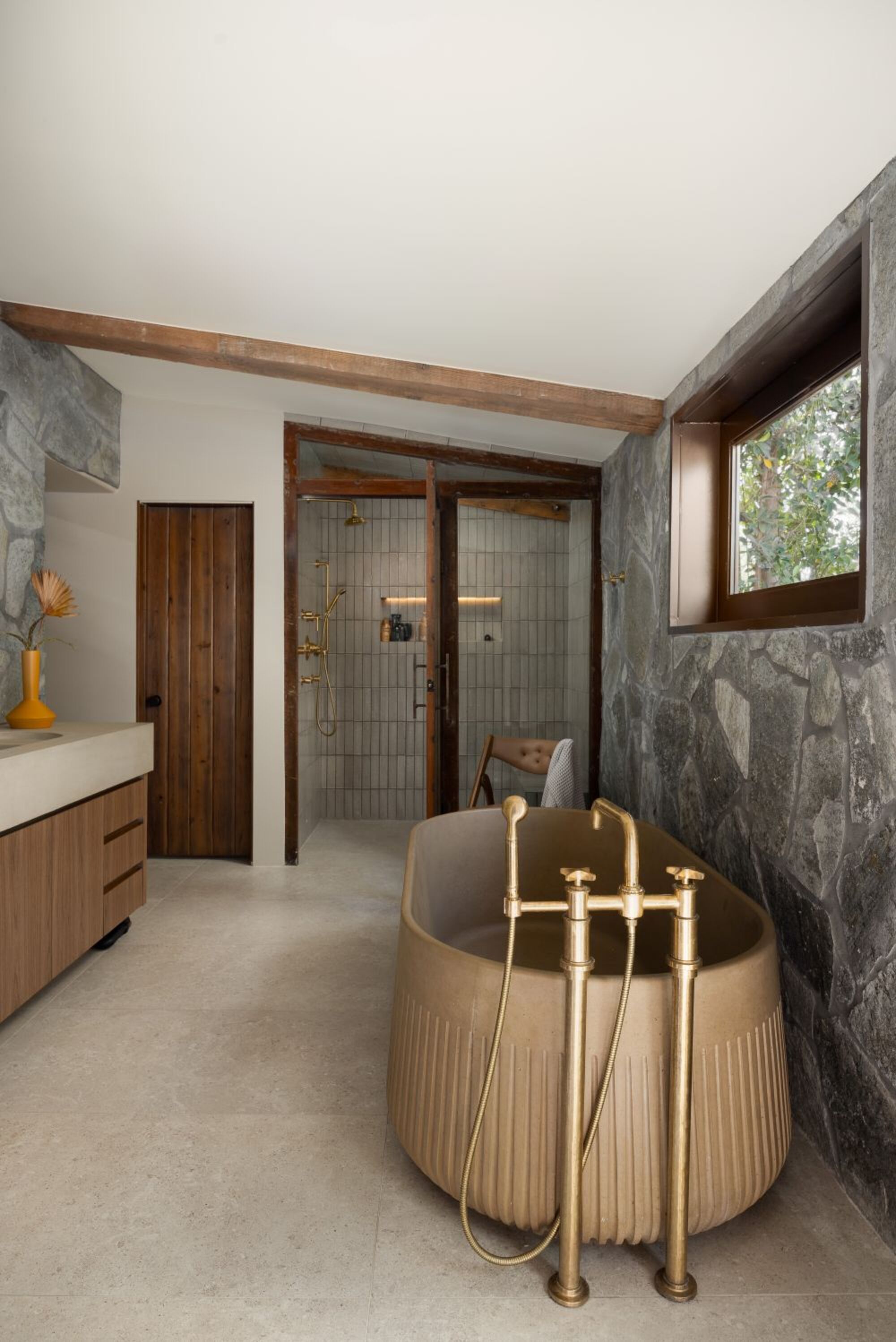 mid century modern bathroom with japandi influence, freestanding taupe textured bath with gold freestanding taps, stone feature wall, tiled shower enclosure, and timber vanity with cream counter