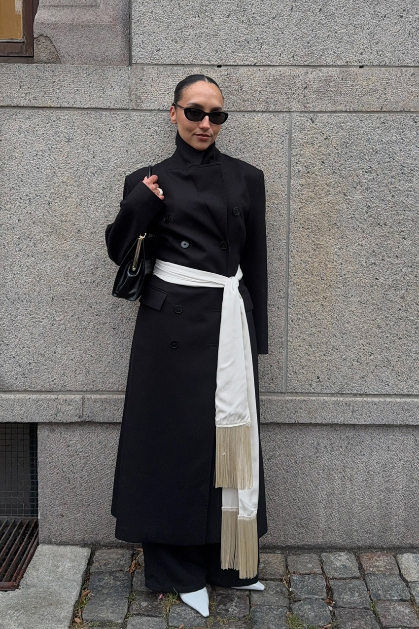 Layering staples for spring 2026 is shown in a street-style photo of a woman standing outside, wearing a long black coat and a white silk scarf cinched at the waist. The coat is layered over a black turtleneck and black wide-leg trousers styled with white pumps, a black shoulder bag, and black sunglasses