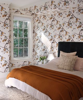 A bedroom with floral wallpaper, a dark orange blanket, and natural light