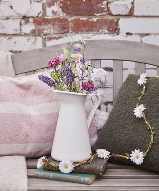 A distressed gray wooden bench with pink and white striped cushion and a gray cushion, with a white vase of pink and purple flowers, in front of a brick wall