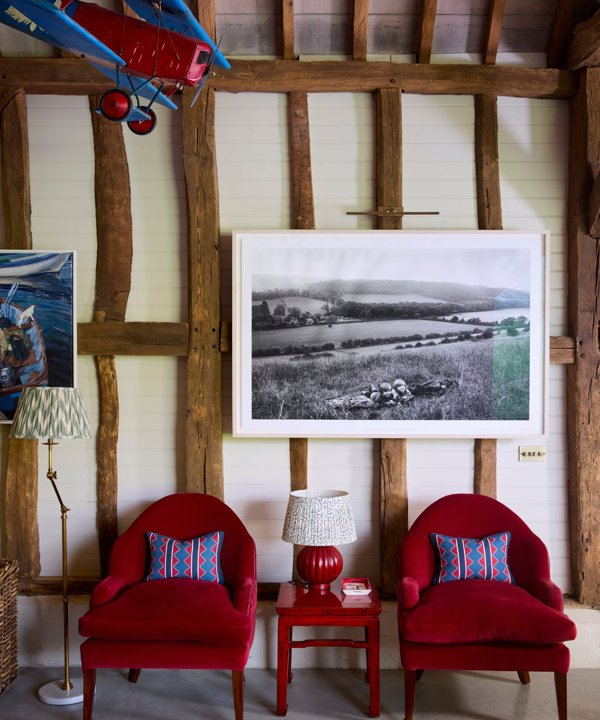 a rustic beamed barn conversation with a red and blue model plane hanging from the ceiling, a black and white landscape framed photo, and two red armchairs