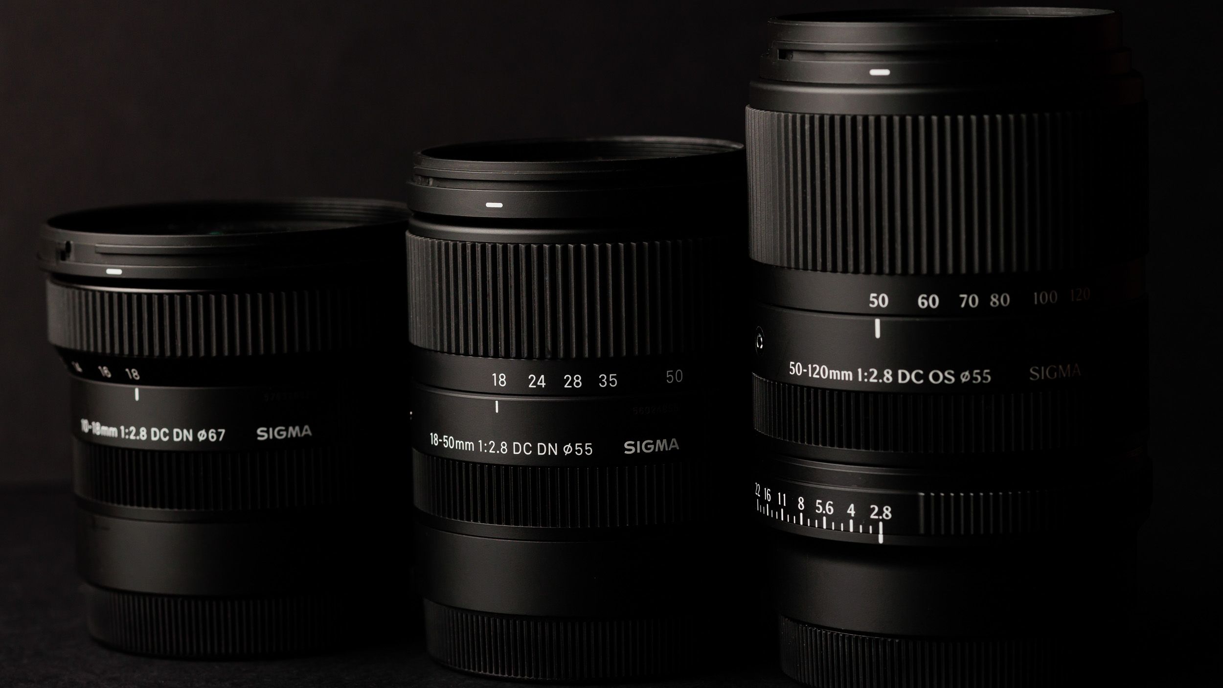 Sigma 50-120mm F2.8 DC OS Contemporary lens alongside two other f/2.8 zoom lenses, in a lowkey lit studio with dark background