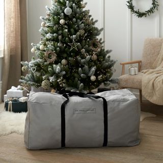 The White Company grey artificial Christmas tree storage bag in front of a Christmas tree with warm white lights