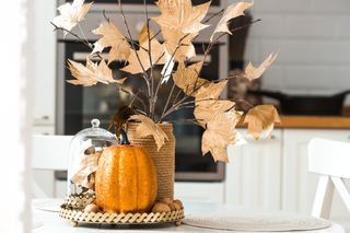 A pumpkin decoration, with a wicker vase displaying some faux gold leaves in a white kitchen.