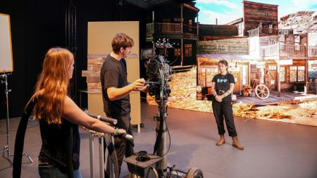 A close-up view of three people working around a camera rig on a dolly in a film studio, with the LED screen backdrop showing the Wild West town.