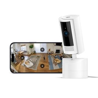 The Ring Pan-Tilt Indoor Cam sits facing slightly down and to the left in front of a smartphone showing a live feed on screen.