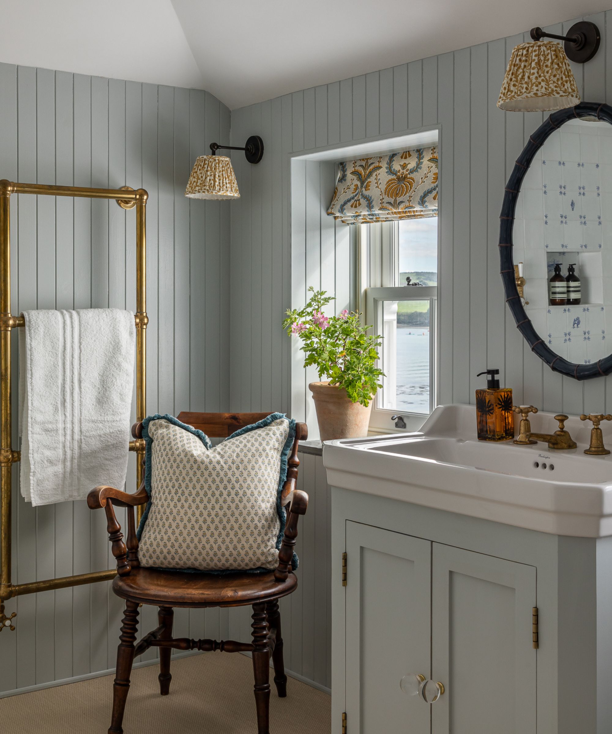 Pale blue painted bathroom with vanity unit, vintage chair with floral cushion, gold towel rack, yellow floral sconces and window with potted plant
