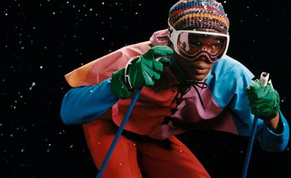 person skiing with a black starry background, dressed in colourful ski gear included cape, beanie, gloves and goggles
