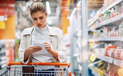 Closeup front view of late 20's woman at a local supermarket checking her shopping list while going through aisles. There are many unrecognizable products on the shelves next to her and in ba