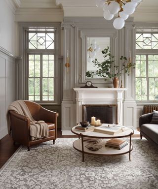 A gray living room with high ceilings and floor-to-ceiling windows, a white fireplace, a leather armchair and a blue and white patterned area rug