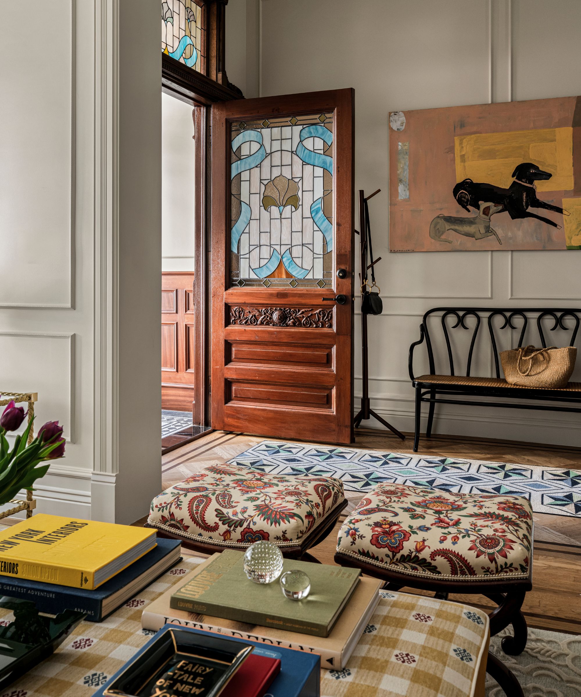 a new york brownstone entryway with mahogany front door with stained glass panels leading into an open plan entry with a bench, artwork of dogs, and a small sitting area with patterned ottomans
