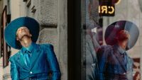 Person in studded blue suit and wide-brim hat leaning against a textured stone wall with their reflection in a nearby glass window