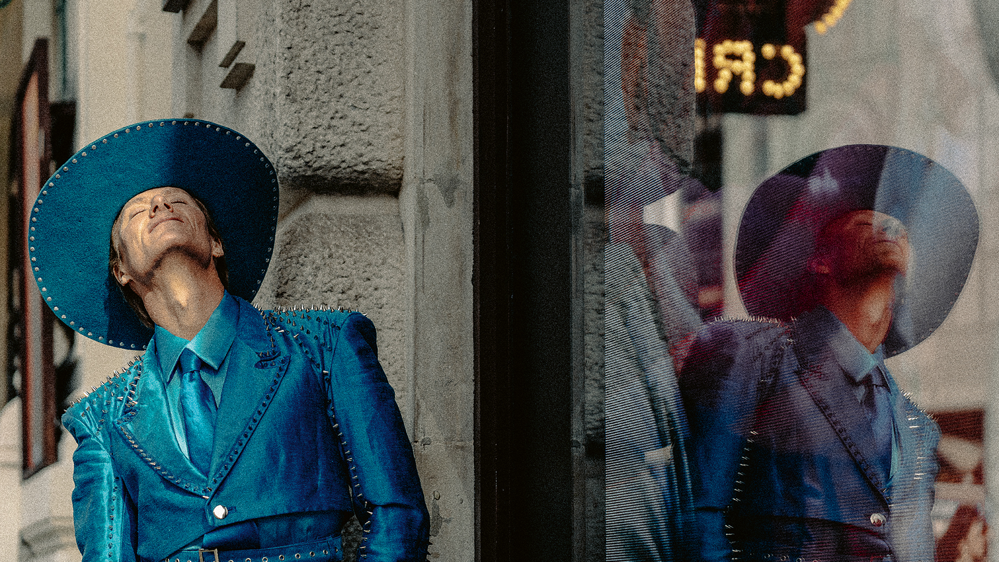 Person in studded blue suit and wide-brim hat leaning against a textured stone wall with their reflection in a nearby glass window