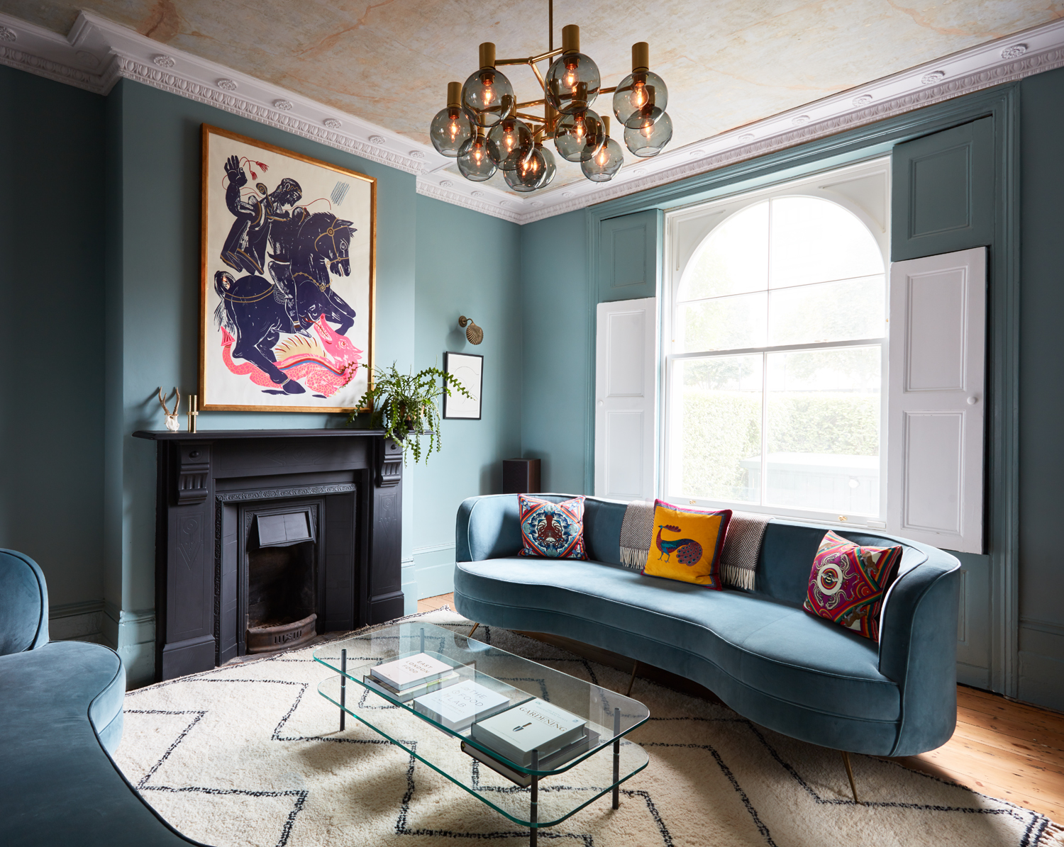 blue living room with blue walls, blue curved sofas, an original black fireplace, glass table, patterned rug, artwork over fireplace, and pendant light