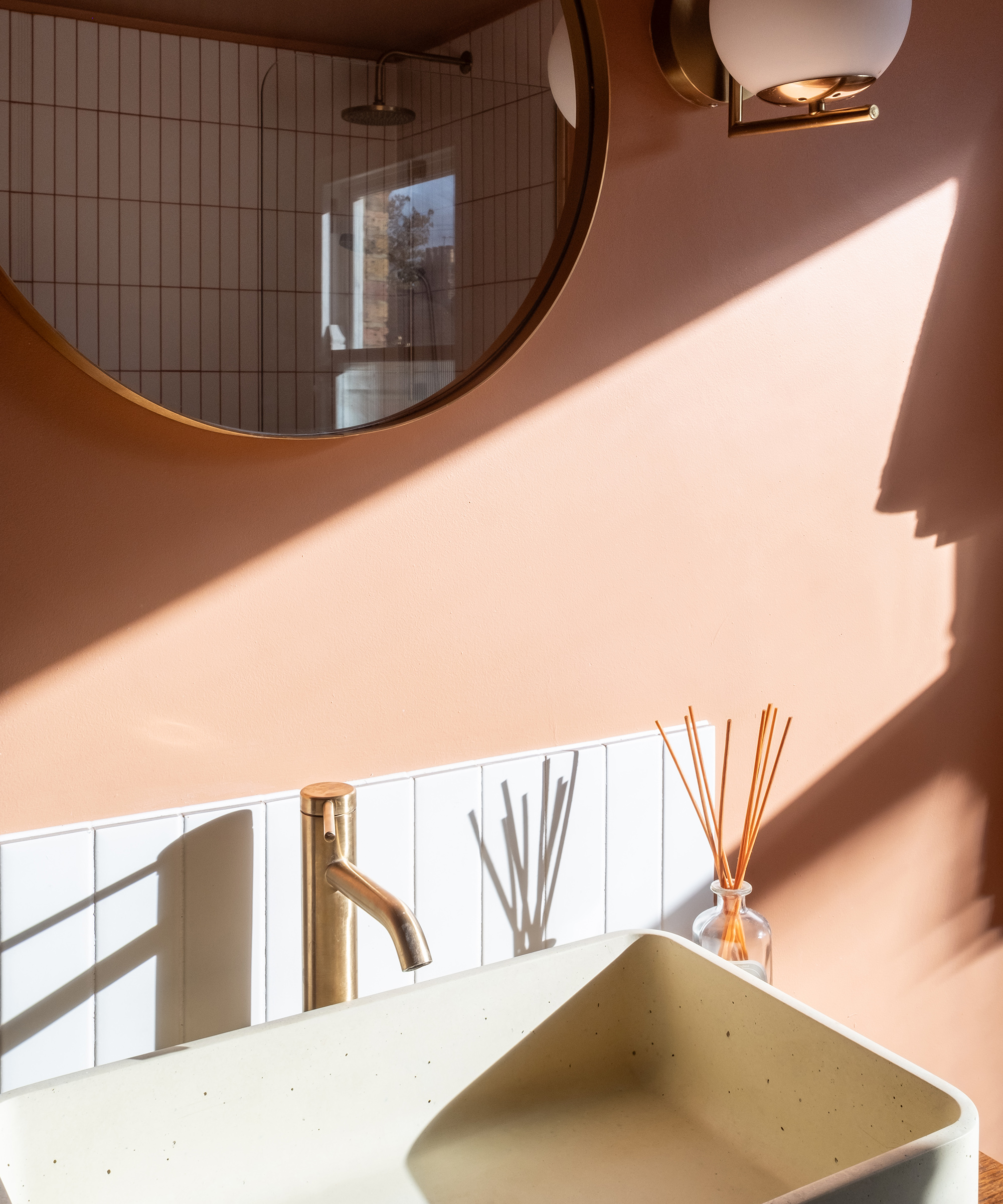 An artful shot of a bathroom sink with playful light and shadow play across the wall. There is a brass tap and chrome framed circular wall mirror
