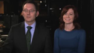 From left to right: Michael Emerson talking and standing next to Carrie Preston who is smiling in a behind the scenes video for Person of Interest posted on CBS's YouTube channel.