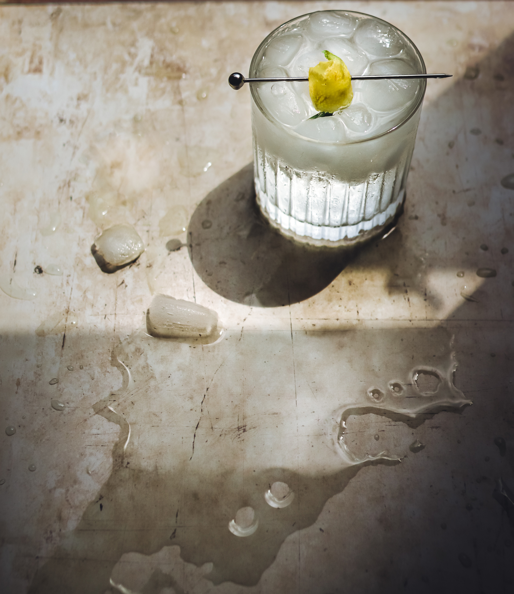 A refreshing cocktail with ice and a lemon garnish rests on a stained surface, surrounded by melting ice and droplets