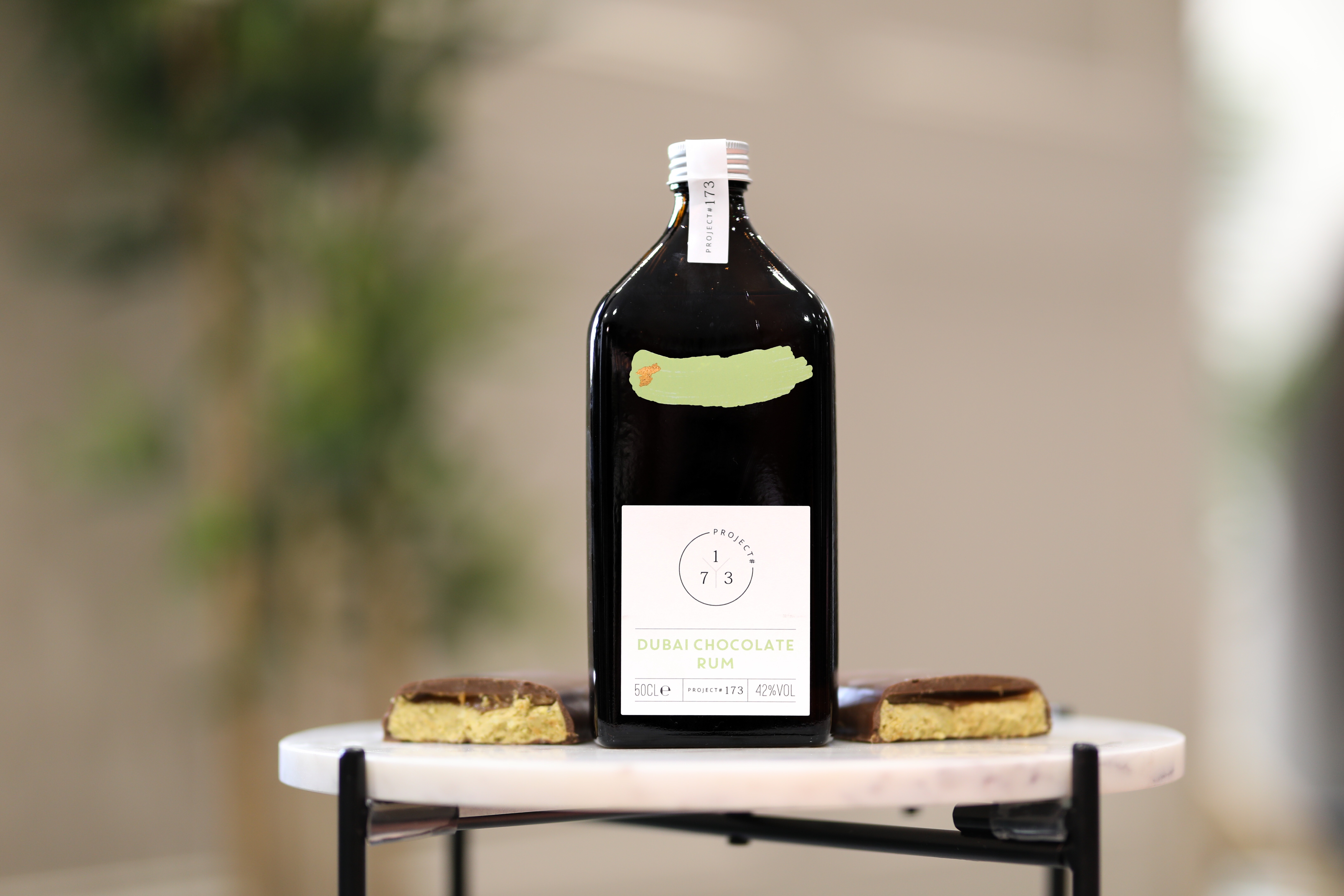 an image of the dubai chocolate rum on a small marble table. There are two pieces of the dubai style chocolate bar either side of the bottle