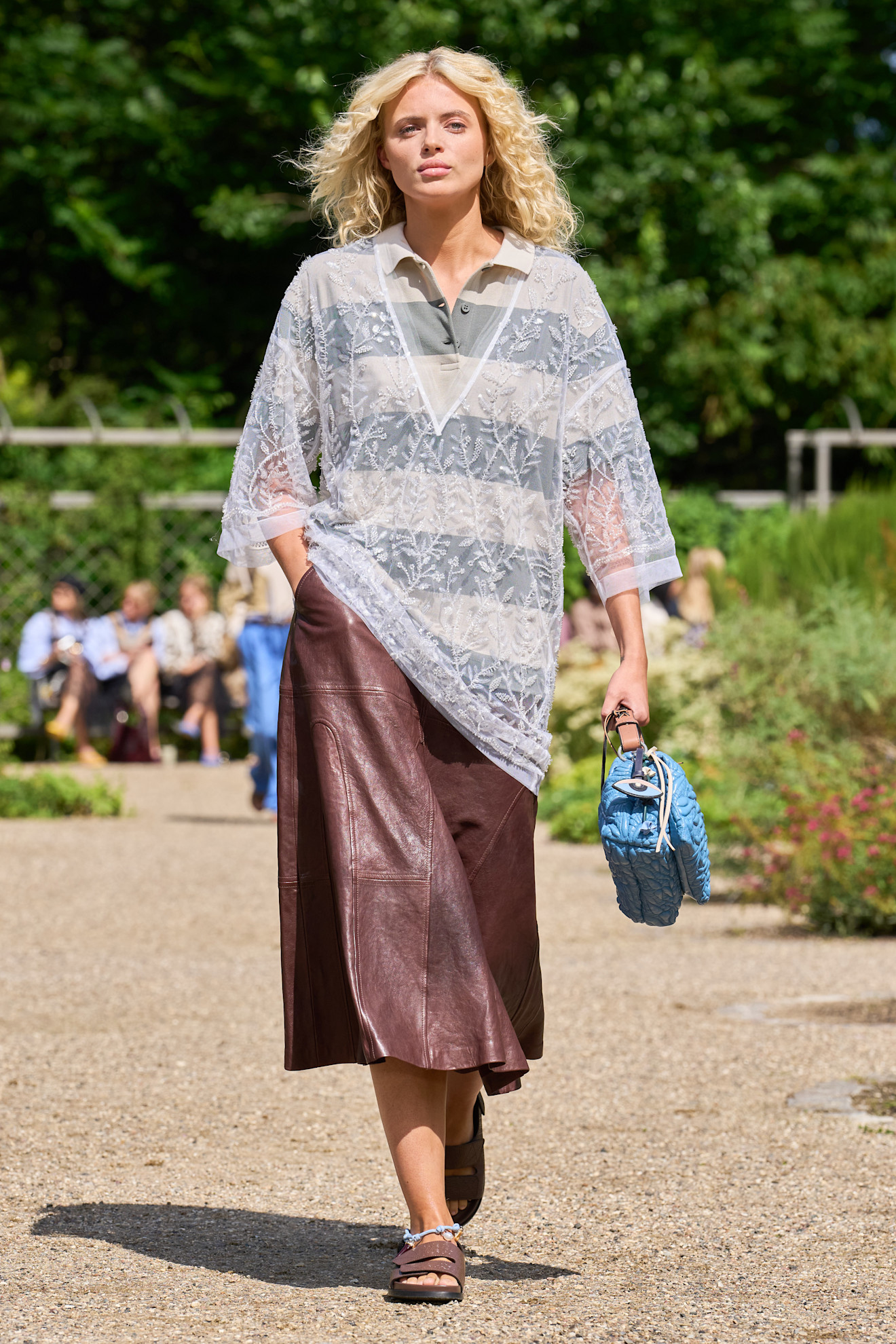 Layering staples for spring 2026 is shown in a photo from Munthe's runway show, with a model walking while wearing an oversized gray striped polo shirt with a sheer embroidered V-neck top layered on top. The look is styled with a brown leather midi skirt, brown flat sandals, and a blue handbag