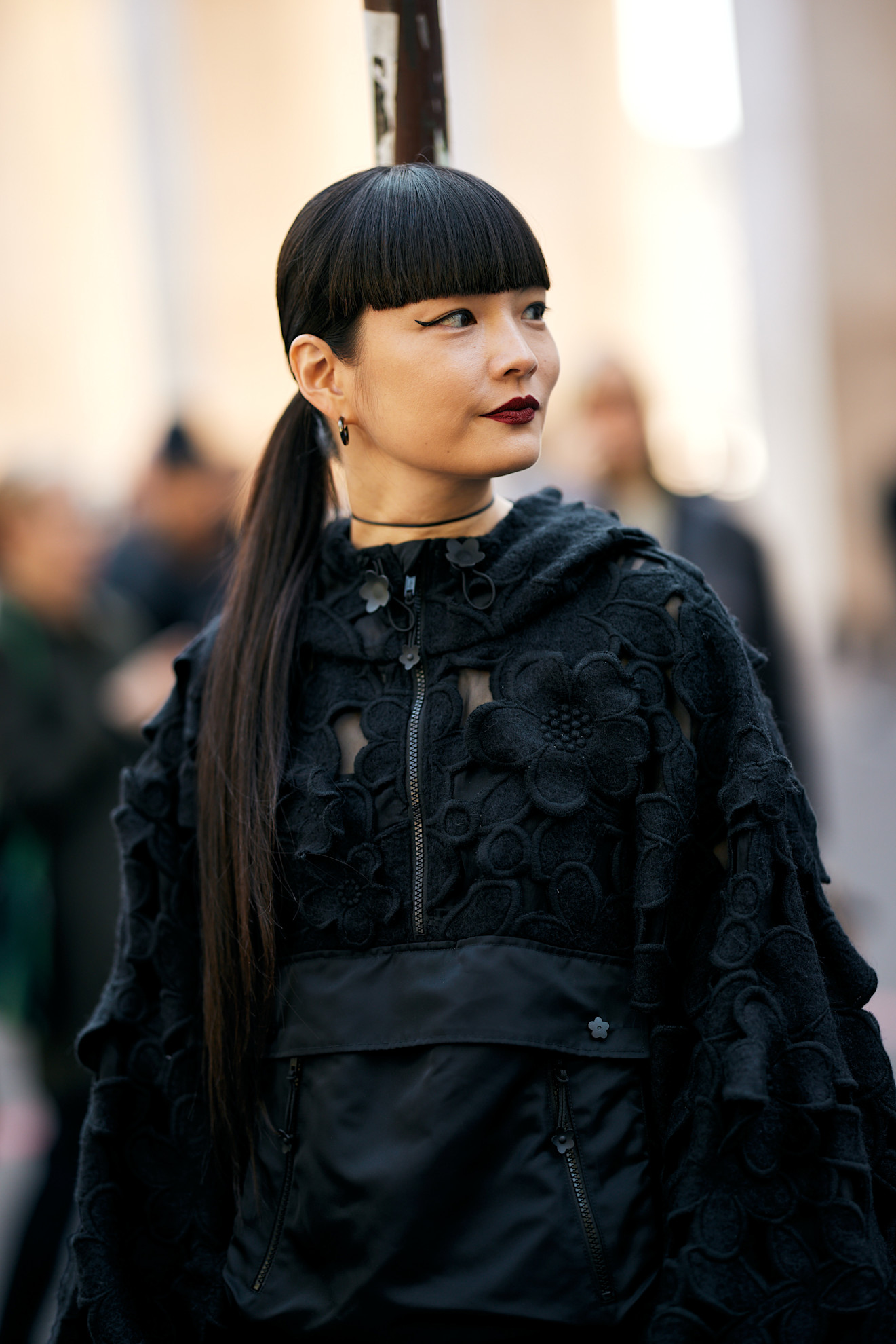a woman in a black lace jacket with a black hair ponytail, blunt bangs, and dark lipstick
