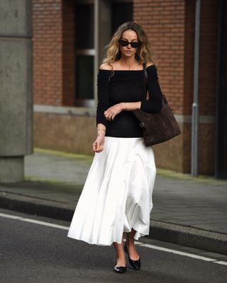 fashion influencer Anouk Yve walking across the street wearing black cat eye sunglasses, a black off-the-shoulder top, brown suede bag, white full skirt, and black flats