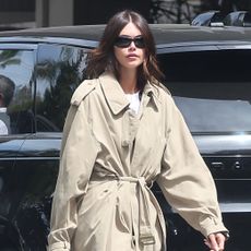Kaia Gerber wearing a trench coat, a white T-shirt, navy satin pants, and red ballet flats