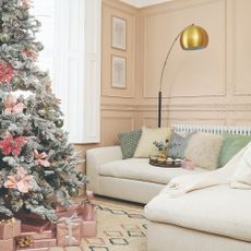 A pastel peach-painted living room with a corner cream sofa and a snowy Christmas tree decorated with pink flowers