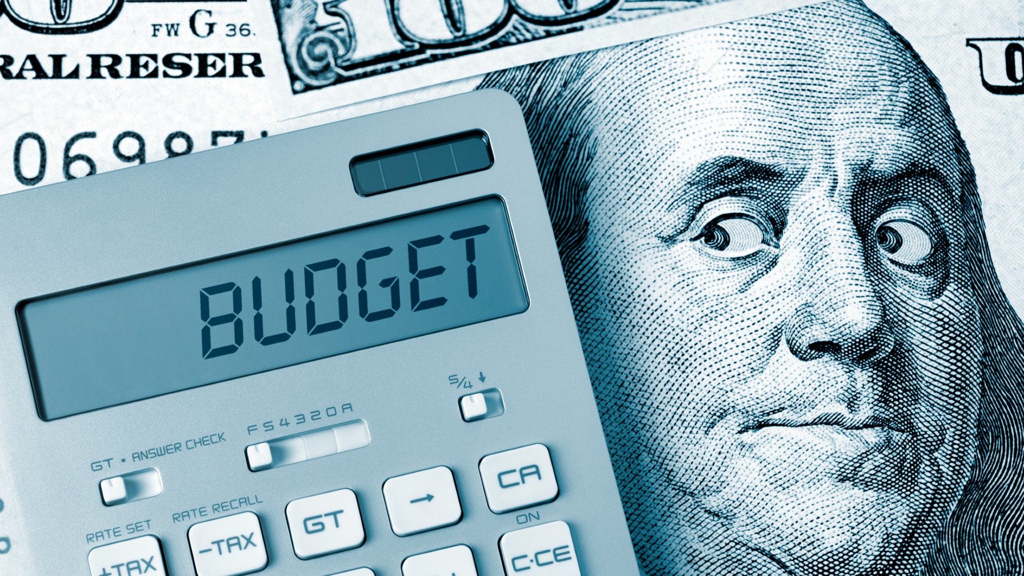 A calculator that says "budget" sitting on top of a hundred dollar bill. Benjamin Franklin's eyes are looking suspiciously at the calculator.