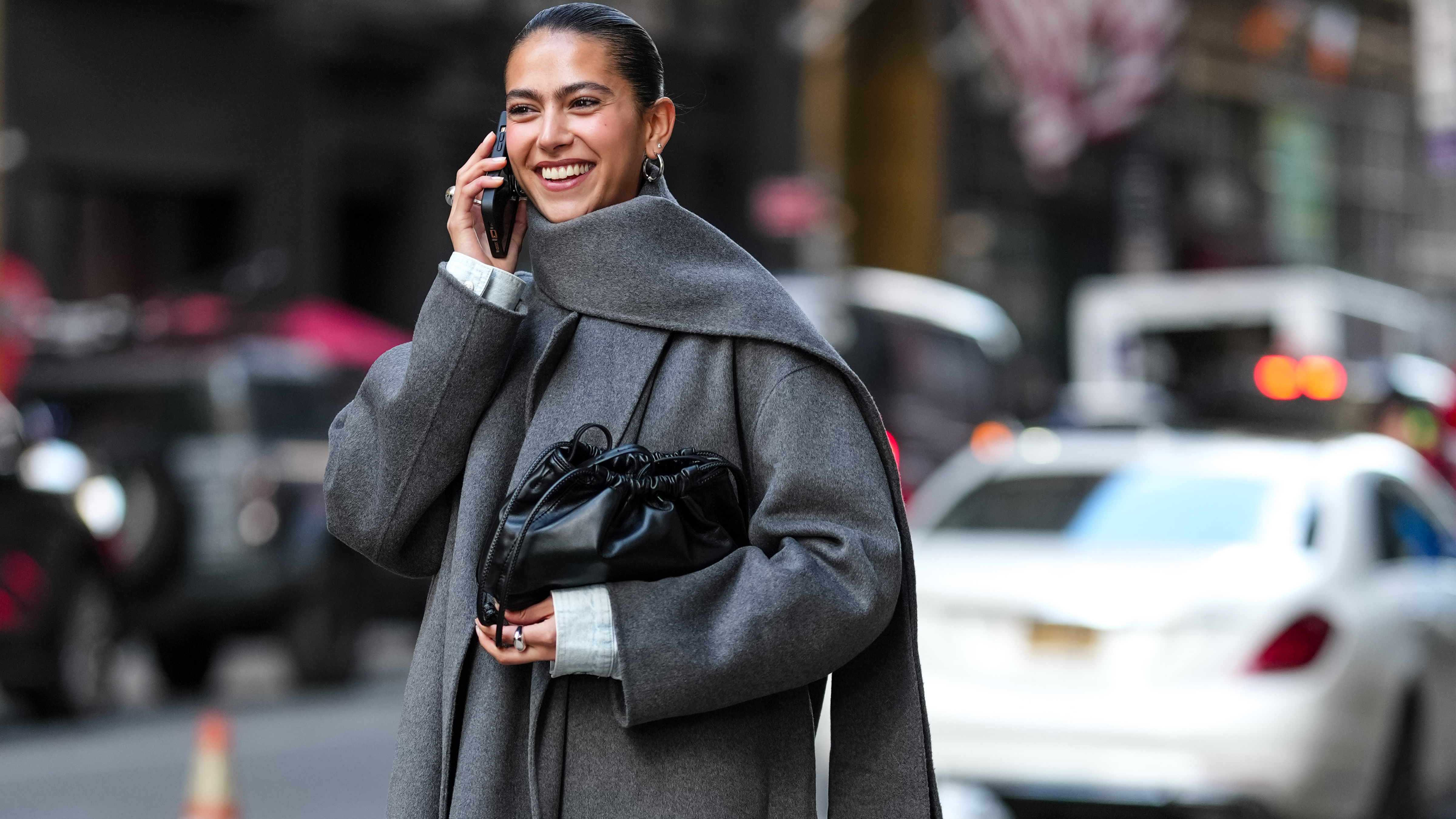 Woman wears a long grey coat with a matching scarf, black leather clutch, knee-high boots, and a denim skirt. 