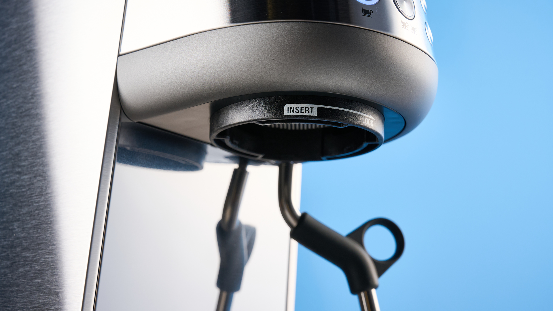 the breville bambino / sage bambino in silver photographed against a blue background with a wooden feature wall, showing the steam wand, group head, water tank, button controls, and espresso
