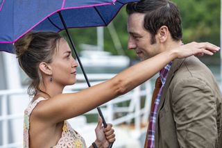 Chelsea Edge and Ralf Little in character as Sophie and Neville in Death In Paradise. They are looking into each other's eyes and smiling. Sophie has one arm resting on Neville's shoulder, and is holding up an umbrella with her other hand.
