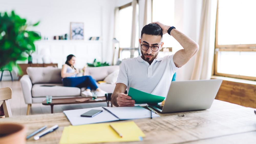 Man rubs head while looking at document. He is in a living room, sitting at a desk with laptop open