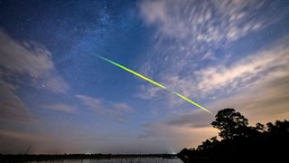 a streak of green light zooms across the night sky above a lake