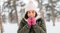 A young woman is shown trying to warm up her hands as she stands in a snowy forest. She is wearing a winter jacket, gloves and a hat. 