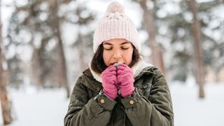 A young woman is shown trying to warm up her hands as she stands in a snowy forest. She is wearing a winter jacket, gloves and a hat. 