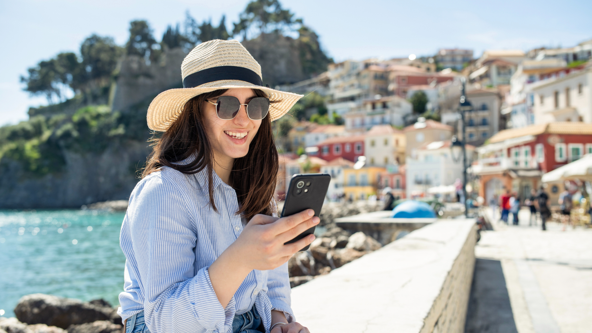A person, sat looking at their phone, while sat on a wall near water in a sunny location.