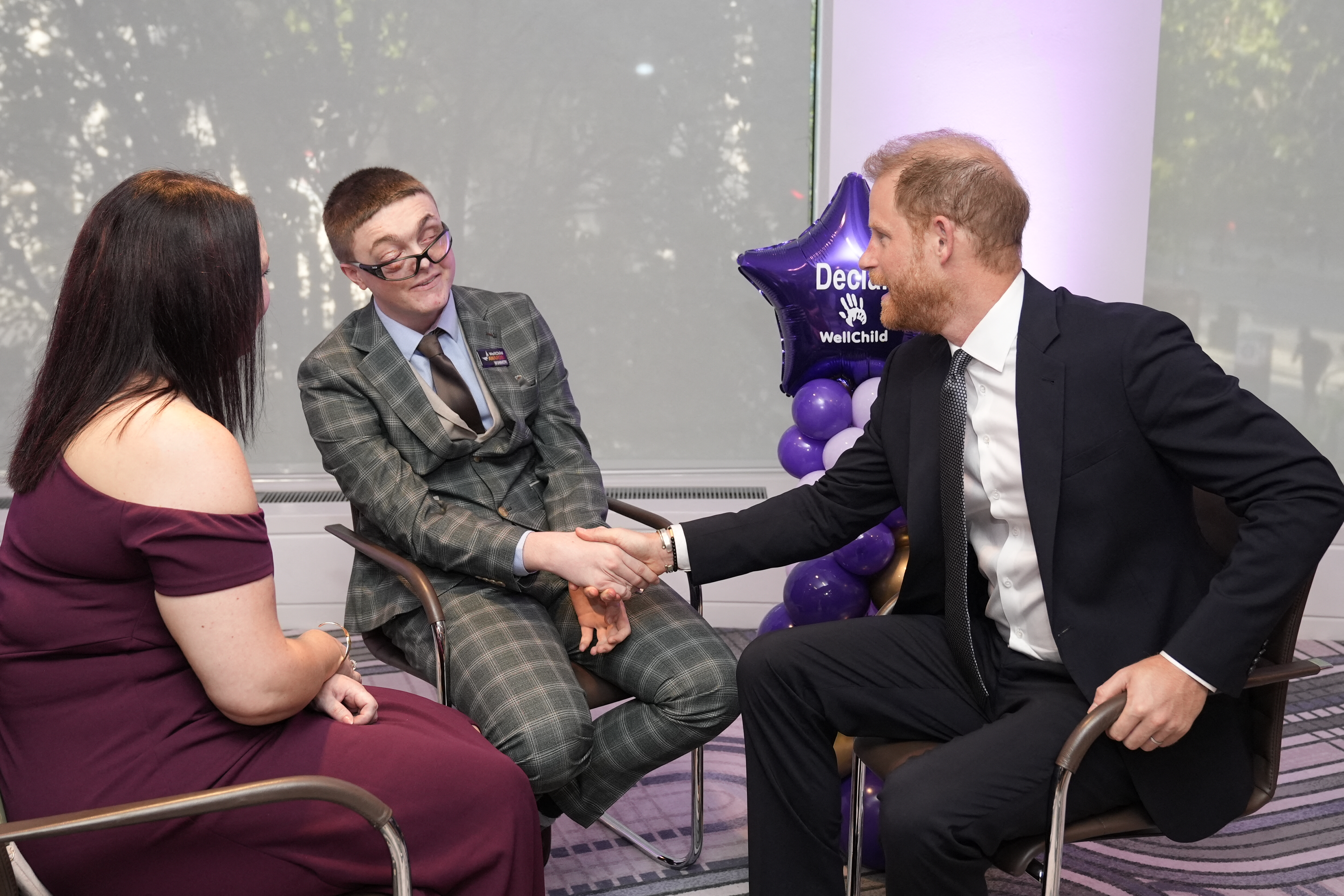 Prince Harry sitting in a chair shaking hands with a young man in a plaid suit