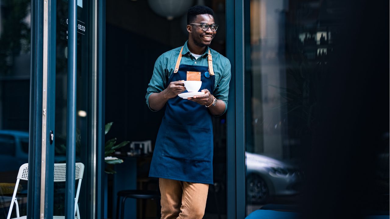 A small business owner smiles and looks relaxed as he stands outside his shop and drinks coffee.