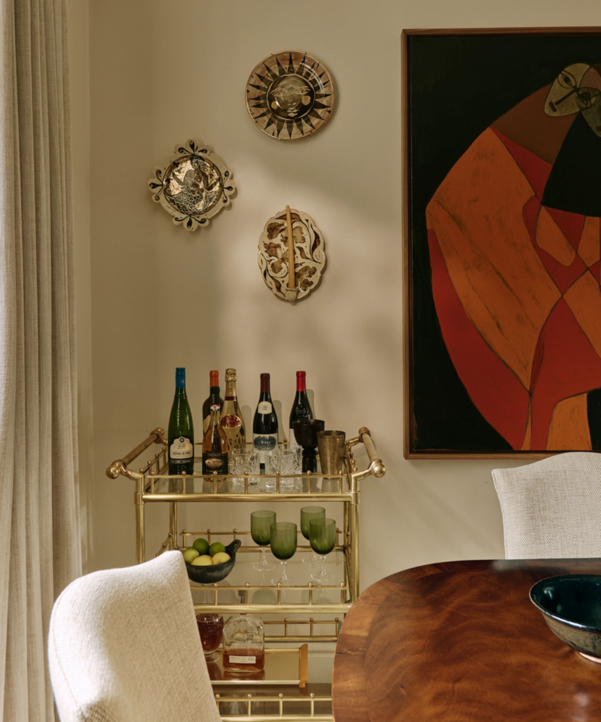 an image of the corner of a dining room painted in warm neutral tones with a brass bar cart, three decorated wall plates and large abstract piece of art