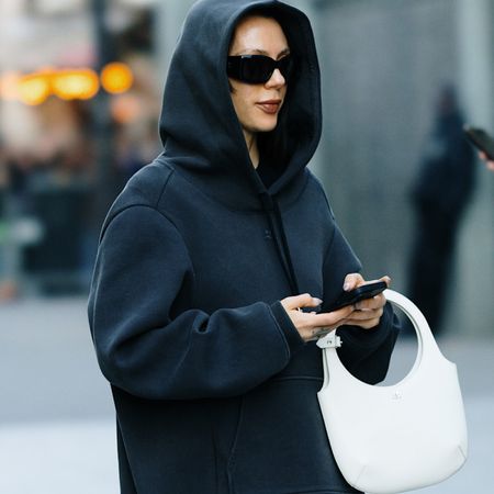 Fashion week attendee wearing black hoodie, black sunglasses, and white handbag