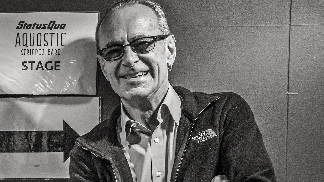 Portrait of Status Quo&#039;s Francis Rossi backstage, next to a sign on the wall that says &#039;Status Quo Aquostic Stripped Bare&#039;