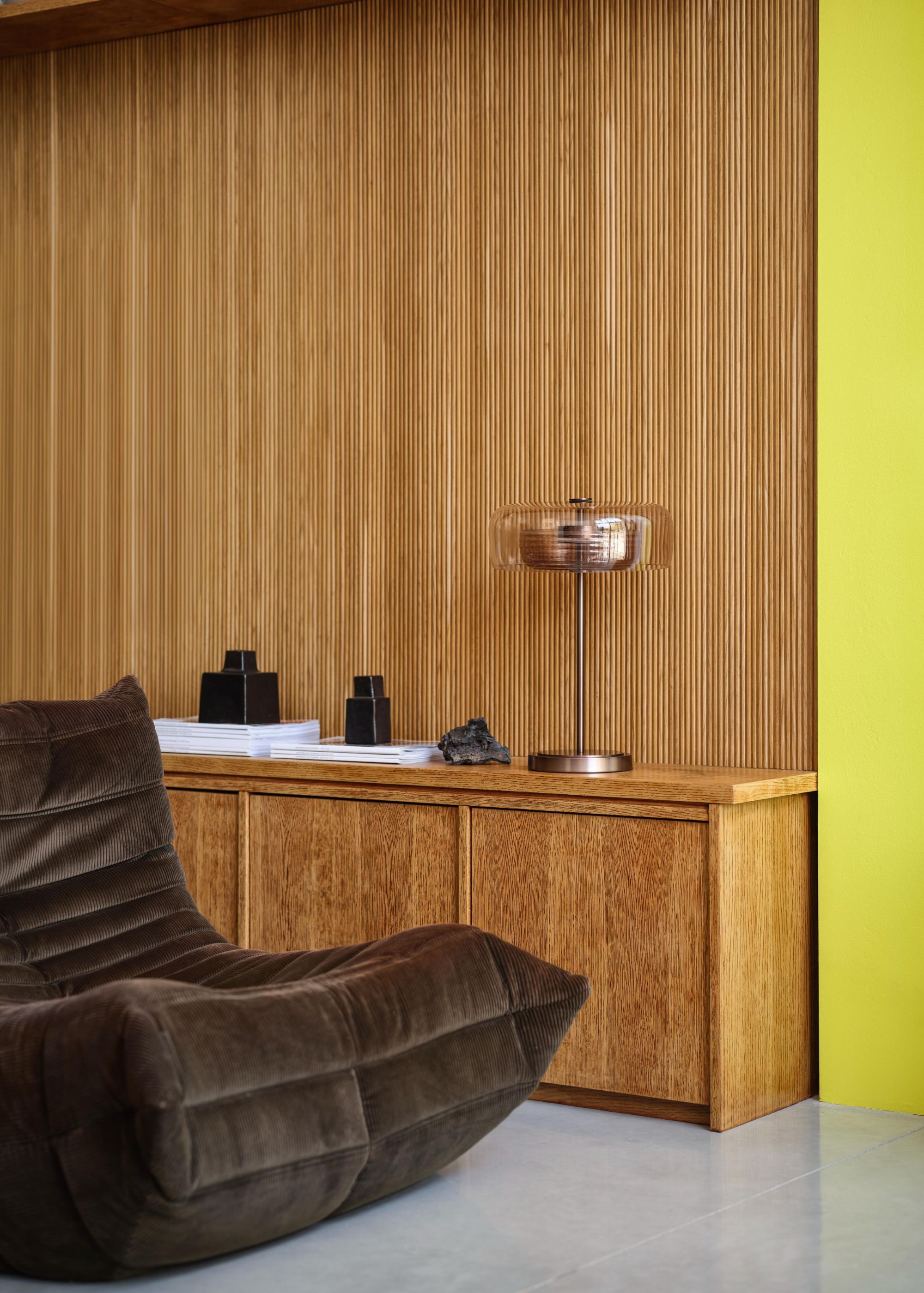 A wood-clad wall with a wooden sideboard in front, a bright yellow wall in the corner, and a slouchy, chocolate brown chair.