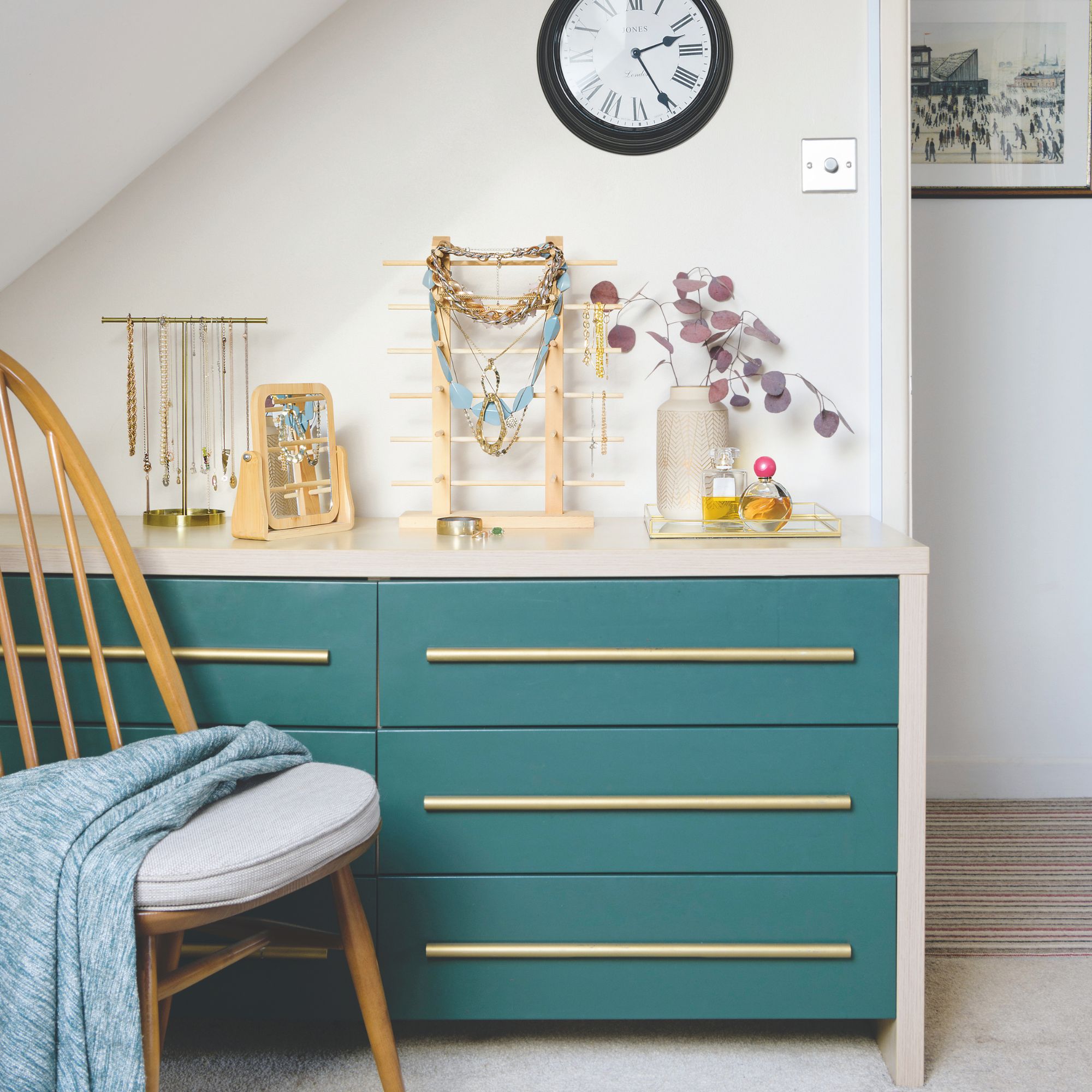 Teal coloured drawers in a bedroom area, topped with accessory holders and a vase, next to a wooden chair