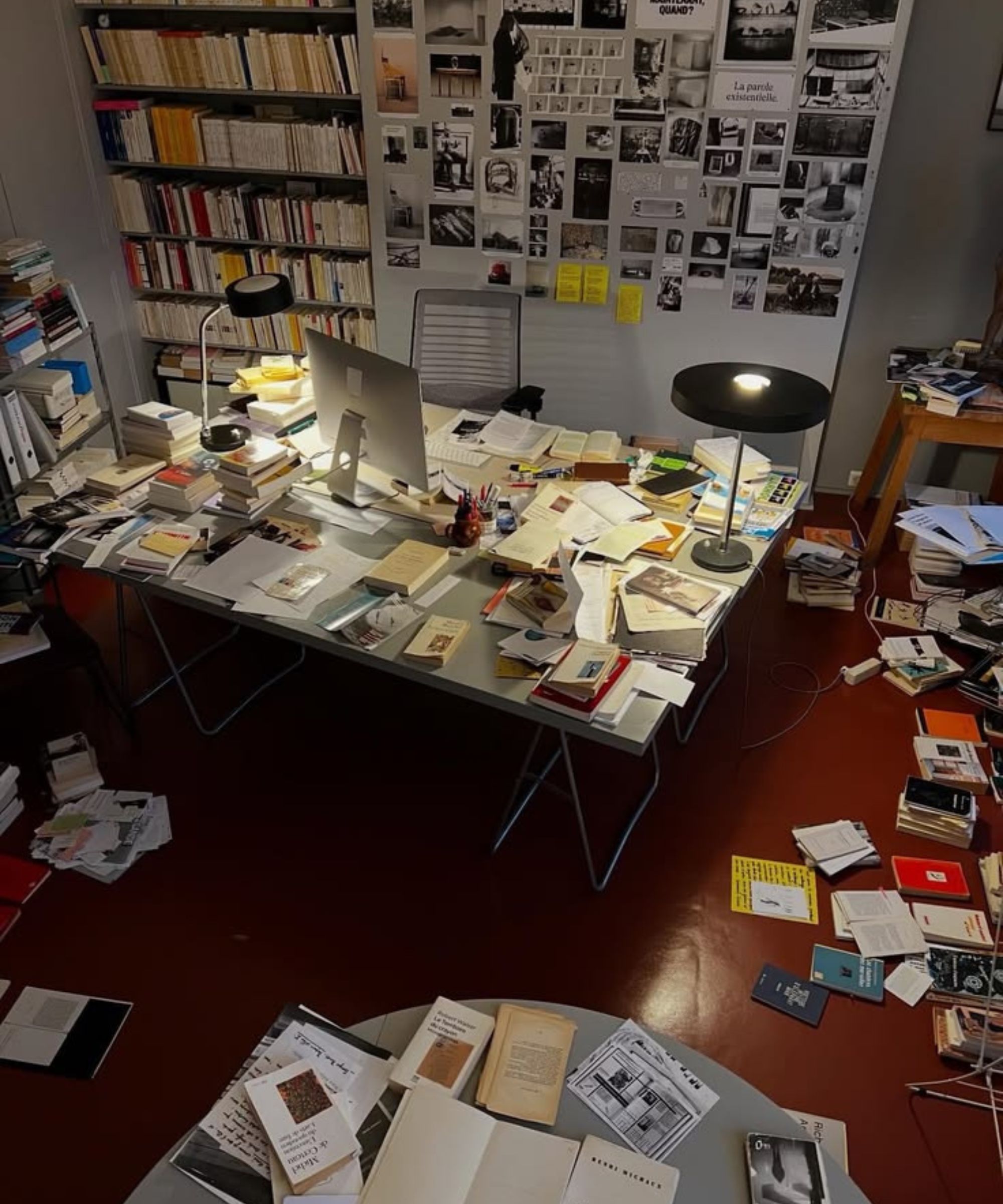 Modern office with large desk covered in books and papers, with computer and lamps. Notice board and bookshelf in background, books and papers on floor