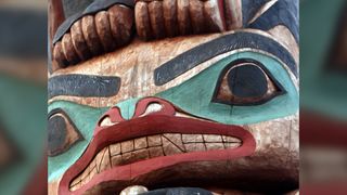 A close-up of a wooden Tlingit totem pole. It has 2 large eyes, heavy black eyebrows, a snort snout complete with red lips and fanged teeth.